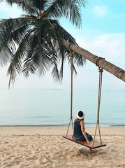 Fototapeta na wymiar Woman sitting on swing hanging on palm tree and looking at calm blue ocean on paradise sandy beach