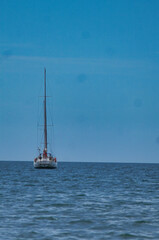 Fototapeta premium The image depicts a sailboat on the open sea under a clear blue sky. The main subject of the image is a white sailboat with a tall mast, floating on the calm sea. The sea has gentle waves and reflects
