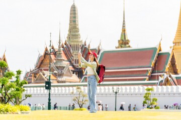 Young asian woman traveler with phone capturing moments and exploring Wat Phra Kaew and The Grand Palace in a happy and casual style during a sunny day