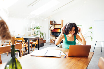 Happy young African American woman in haedphones is sitting working on a laptop at home. Remote...