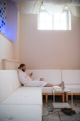 Man with beard and hair pulled back to ponytail relaxing in a plush white robe on a white couch using smart phone