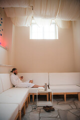 Man with beard and hair pulled back to ponytail relaxing in a plush white robe on a white couch...