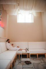 Man with beard and hair pulled back to ponytail relaxing in a plush white robe on a white couch using smart phone