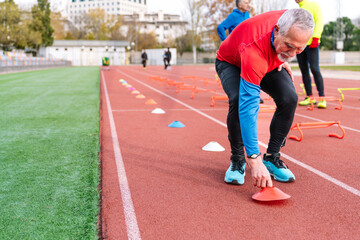 Focused elderly athlete in red shirt setting up agility cones on a sports track with peers in background.