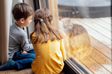 Cute children in casual clothes are sitting on the floor near the panoramic window in the living...