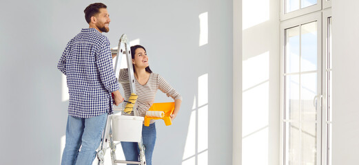 Smiling happy couple painting the wall of their new home holding paint rollers and looking at the window standing on the ladder. Married man and woman doing repair renovation. Banner.