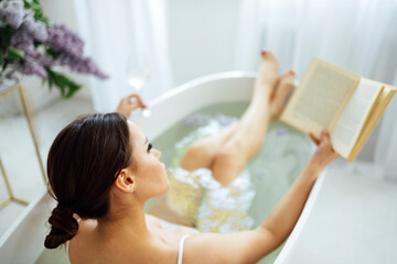 young brunette woman reading the book and drinking white wine in bath