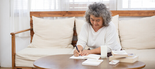 Asian senior woman sitting on couch reading, writing or working in living room at home. Doing financial document. retired people living lifestyle insurance concept