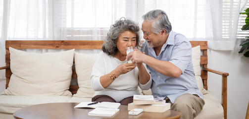 Asian senior man help and support his wife to drinking milk sitting on couch in the living room....