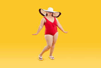 Full length studio portrait of happy beautiful young fat plump woman wearing red one piece swimsuit and big wide holiday beach sun hat that covers her eyes posing isolated on yellow color background