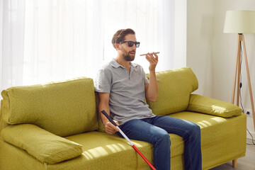 Portrait of a young bearded blind man in black eyeglasses holding cane and using digital assistant on mobile phone sitting on sofa at home. Visually impaired person talking on mobile phone.