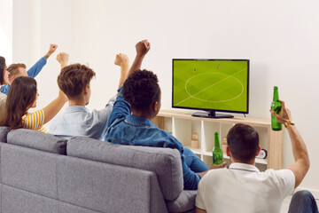 Group of happy diverse friends watching soccer on TV. Young multiracial people sitting on sofa, looking at television screen, watching football, drinking beer, raising hands up and cheering. Back view - obrazy, fototapety, plakaty