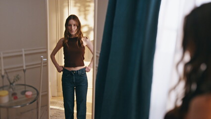 Woman reflection trying clothes large jeans at home. Smiling girl getting ready