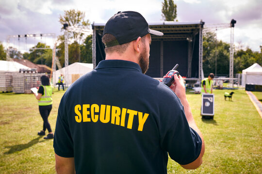 Rear View Of Security Team At Outdoor Stage For Music Festival Or Concert Talking Into Radio