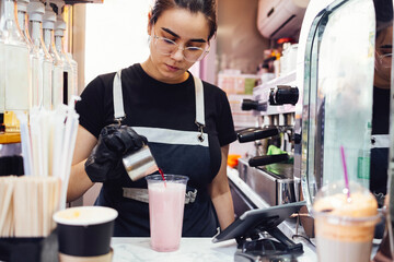 Young woman in black gloves prepares milky pink drink in street takeaway cafe