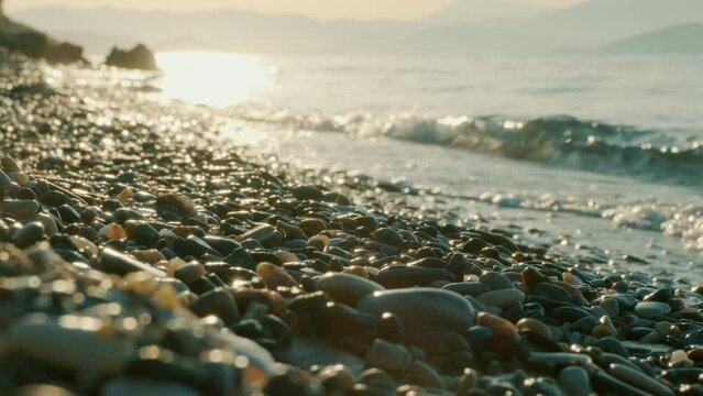 A perfect beach scene. Waves gently crash into a pebble-strewn shore as the sun sets in slow motion. Holiday concept.