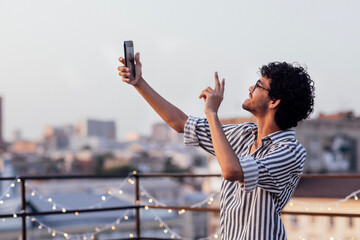 Young curly-haired guy in glasses and striped shirt answers video call on phone. Smiling mixed-race...