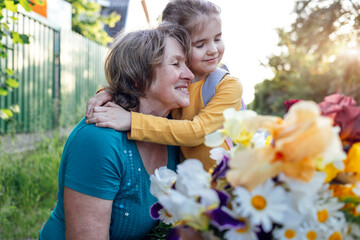 Cute granddaughter hugging her happy grandmother. The hugs of an elderly woman and a little girl against the backdrop of summer flowers, trees and sunset.