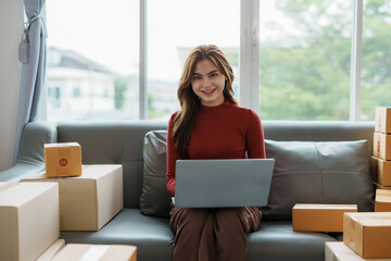 Smiling young business owner woman prepare parcel box and standing check online orders of product for deliver to customer on laptop computer. Shopping Online concept.