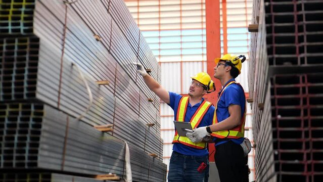 Slow-motion 4K video. Senior engineer wearing safety uniform workwear and holding a tablet instructs or on the job training a trainee engineer in a formed steel pipe storage area in industrial plants.