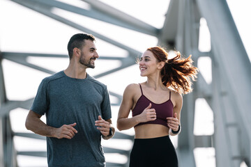 Young adult couple running together in the city looking at each other while jogging. Happy athletic male and female in sportswear training workout outside.