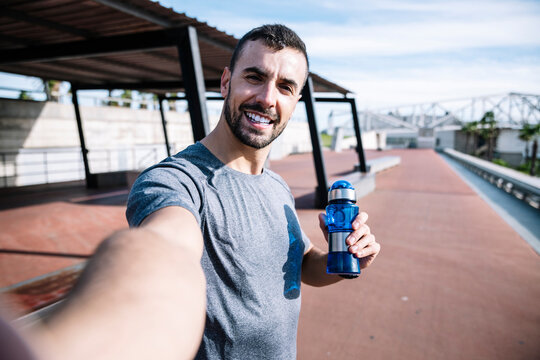 Runner man drinking water and taking a selfie after exercise outside. Handsome sports guy holding a bottle taking a picture and looking at camera.