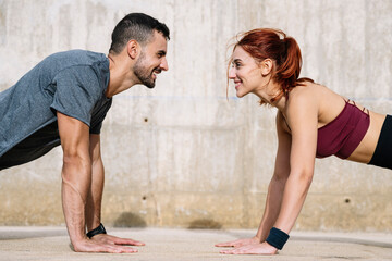 Determined young adult couple exercising in the floor looking each other. Joyful athletic male and female doing push ups in the street in sportswear training workout outside.  - obrazy, fototapety, plakaty