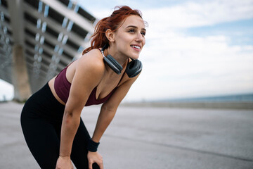 Young smiling fit woman in sportswear resting after run wearing headphones. Athletic female listening music taking a breath during a training workout leaning on her knees after exercising. - obrazy, fototapety, plakaty