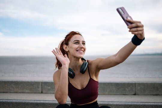 Joyful sports woman taking a selfie wearing headphones sitting outside. Young adult fit athletic female in sportswear taking a picture with smartphone