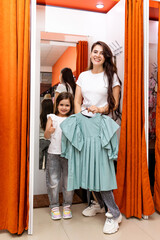 Mother and her little daughter in a fitting room in a shopping mall. Girl trying on a dress before buying. Vertical photo