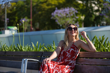 South American woman, young, beautiful, blonde with flower dress and sunglasses, sitting on a wooden bench, smiling and happy. Concept travel, destinations, Europe, Mediterranean, vacation.