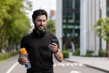 A young sporty man with headphones is standing on a city street, holding a bottle of water and...