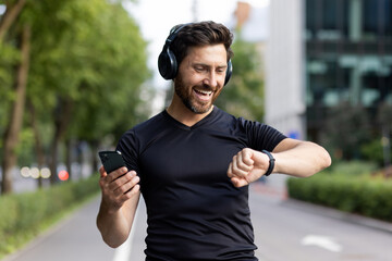 A happy young man is standing on a city street wearing headphones, holding a phone in his hand and looking contentedly at a smart watch. Close-up photo