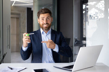 Portrait of a young businessman sitting at a desk in the office, holding and pointing to a cough inhaler - obrazy, fototapety, plakaty
