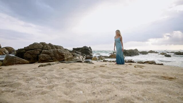 Sun-kissed woman strolls along beach in blue dress, waves crash on rocky shore behind. Leisure walk on sandy coastline, tranquil sea backdrop. Vacation on exotic beach, female enjoying solitude.
