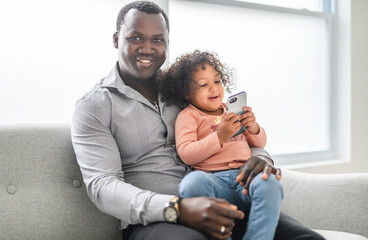 Happy Black American father with little daughter at home using cellphone