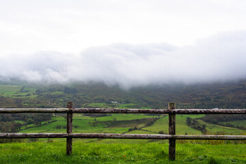 Beautiful landscape seen from the edge of a road with wooden fence of green fields and farms with very low clouds