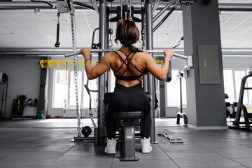 Muscular athletic female bodybuilder in black suit pulled on sports simulator in gym. Back muscles...