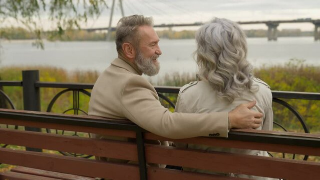 Back view happy senior mature 60s husband wife hugging on bench outdoors healthy retirement couple mid aged woman man talk laugh relax enjoy romantic weekend together love old age in city park nature