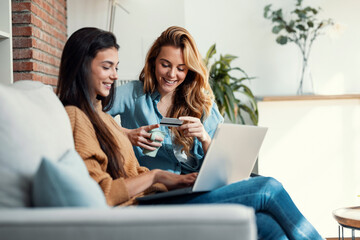 Two happy young women using computer while paying with credit card their vacation sitting on the couch at home. - Powered by Adobe