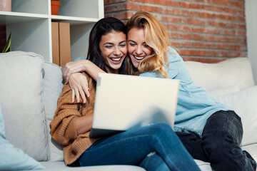 Two happy beautiful women doing a video call with laptop while hugging each other sitting on the...