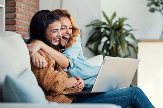 Two happy beautiful women doing a video call with laptop while hugging each other sitting on the couch at home.
