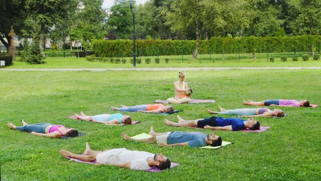 Yoga group relaxes in savasana while woman plays singing bowls, creating serene soundscape in verdant city park, merging meditation and sound healing