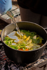 A gloved hand pours white wine into a black pot filled with sliced apples, mandarines, and fresh mint, set on a grill over glowing coals, capturing the essence of outdoor winter cooking