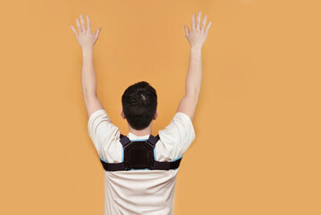 A young guy wears a special posture corrector to keep his back straight and healthy and does special exercises with arms up, rear view. The boy has a corset on his back, back hurts - obrazy, fototapety, plakaty
