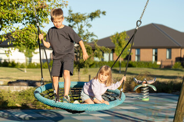 Brother and sister are playing on the playground outside. Children ride on a swing. Happy childhood