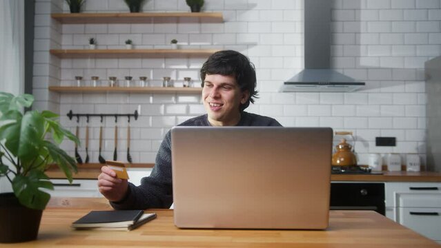 Young man sitting in the kitchen entering credit card number on laptop for makes secure easy distant electronic payment at home
