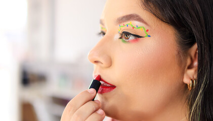 Close-up of a young Latina woman with light brown eyes applying fiery red lipstick as part of her makeup
