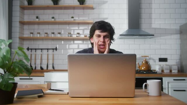 Sad young man sitting against the kitchen using laptop having anxiety and stress. Upset man reacting to loss, bad news