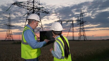 Electricians discuss project data on laptop looking at power transmission lines in field. Engineers plan power distribution plant development on laptop at dusk countryside. Electricity generation